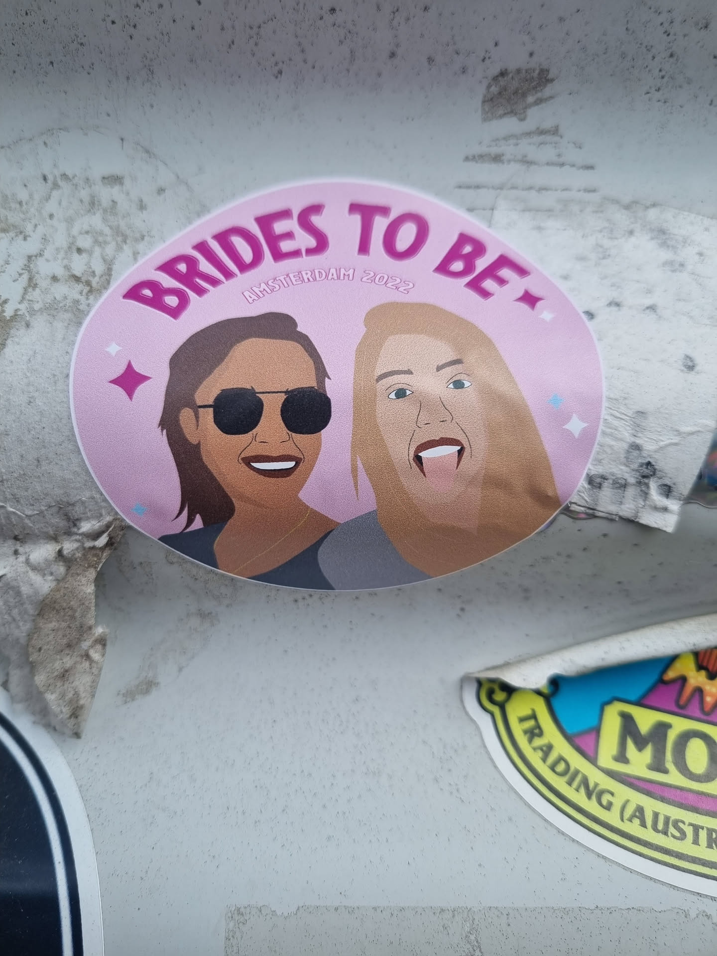 A pink round sticker with an illustration of two women wearing sunglasses, one of them is sticking her tongue out. The text 'Brides to be' is written in a bold purple font, and 'Amsterdam 2022' is written in a smaller font underneath.