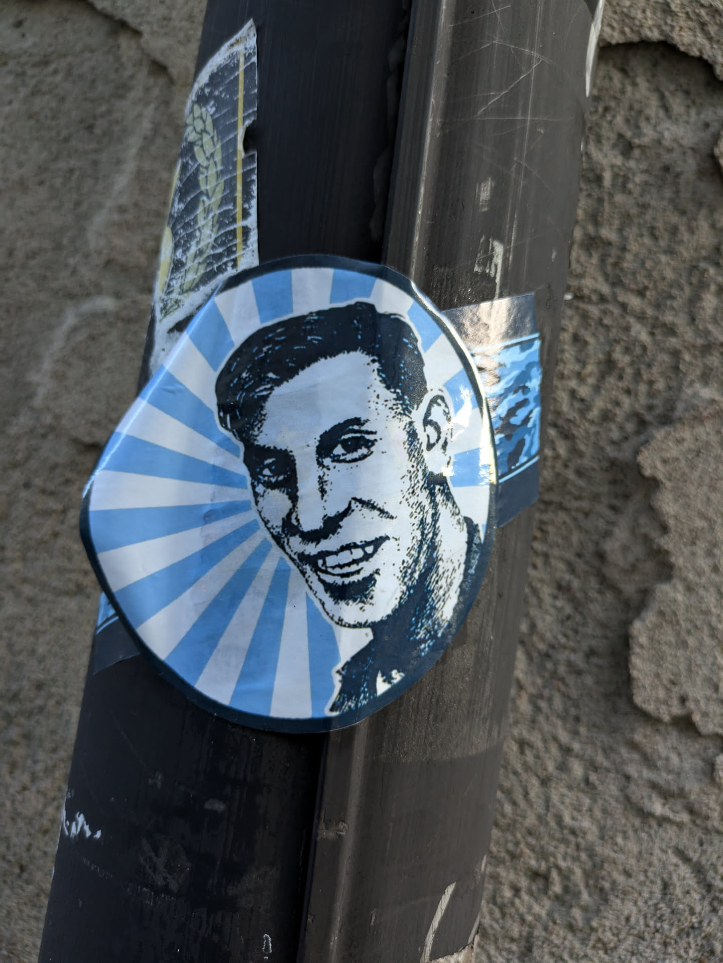 This sticker shows a stylized portrait of a man, done in blue tones against a sunburst background. The style is reminiscent of mid-20th century graphic design.  The sticker is affixed to a dark pole outside.