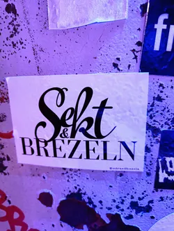 A sticker with the text "Sekt & Brezel" in a stylized font. The background is white, and the text is black.  The sticker appears to be on a wall with other stickers and markings.