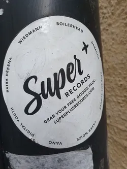 Street sticker Round white sticker with black text and logo.  The main text is "Super Records" in a stylized script font.  Smaller text includes instructions to grab a free goodie pack and a website address: superplusrecords.com. There are also several names around the edge: Wiedmann, Boilerhead, Baiba Dekena, Digital Youth, Vano, Dabadox, and Laser Noize.