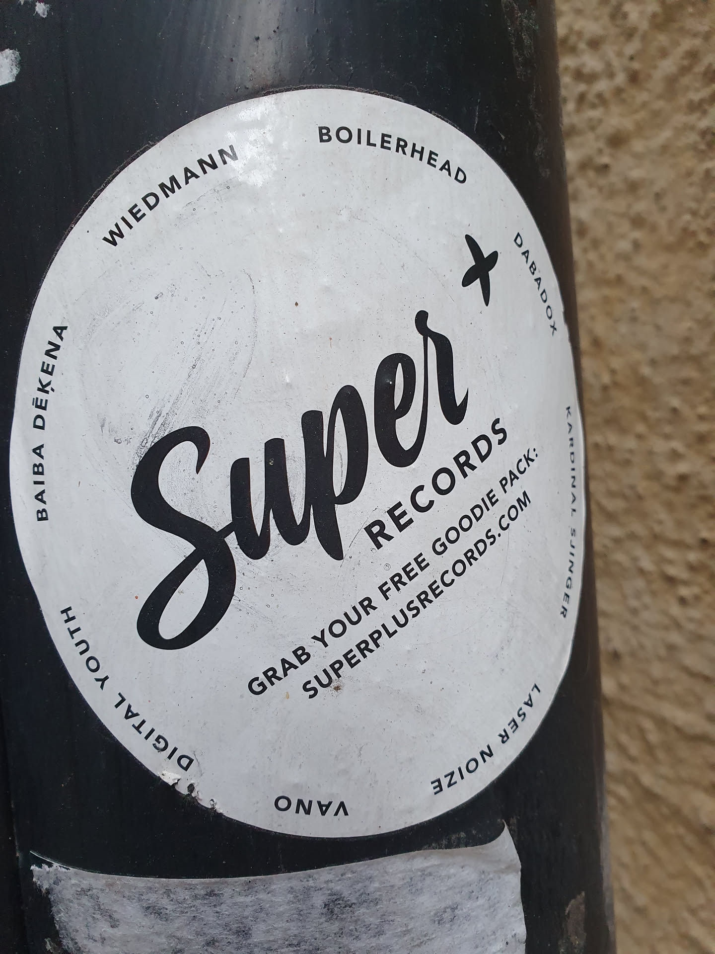Round white sticker with black text and logo.  The main text is "Super Records" in a stylized script font.  Smaller text includes instructions to grab a free goodie pack and a website address: superplusrecords.com. There are also several names around the edge: Wiedmann, Boilerhead, Baiba Dekena, Digital Youth, Vano, Dabadox, and Laser Noize.