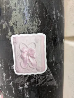 A square sticker depicting a pink, textured design resembling a book cover or decorative box. A pink ribbon with a small rhinestone accent is centrally located. The sticker has a slightly irregular, possibly hand-cut edge and is affixed to a dark-colored, textured surface, possibly a pole or pillar.