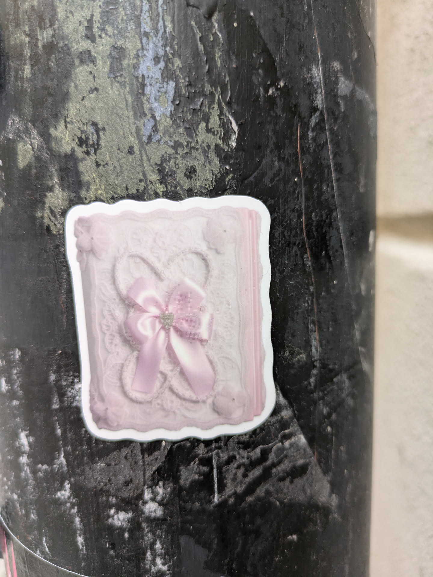 A square sticker depicting a pink, textured design resembling a book cover or decorative box. A pink ribbon with a small rhinestone accent is centrally located. The sticker has a slightly irregular, possibly hand-cut edge and is affixed to a dark-colored, textured surface, possibly a pole or pillar.