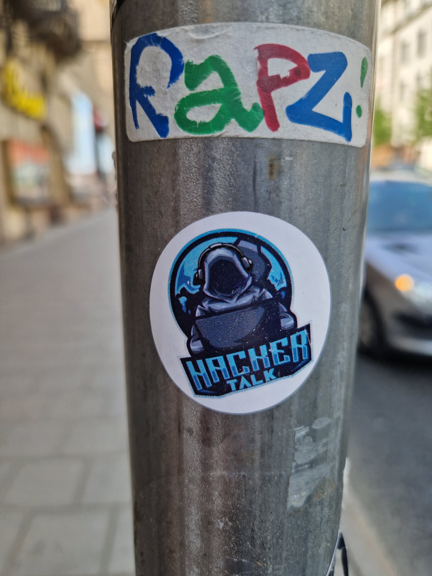 A round sticker featuring a stylized illustration of a hooded figure sitting in front of a laptop, set against a blue background with digital elements. The text "Hacker Talk" is prominently displayed below the illustration. The sticker appears to be professionally printed and is affixed to a metal pole.