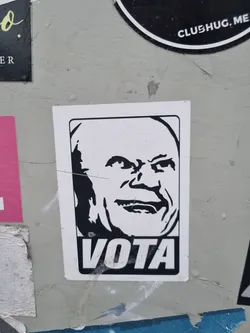 Black and white sticker featuring a stylized portrait of a bald man with a mischievous grin. The word 'VOTA' (Spanish for 'Vote') is printed prominently below the portrait.