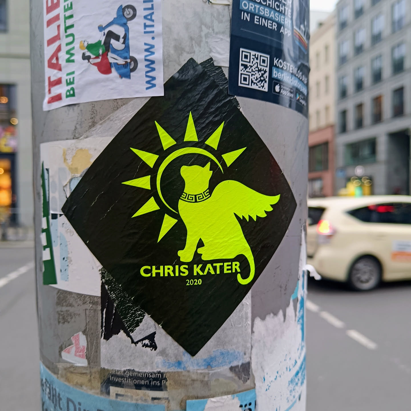 A black diamond-shaped sticker with a yellow cat with wings, surrounded by a sun. The text 'CHRIS KATER 2020' is written below the cat.