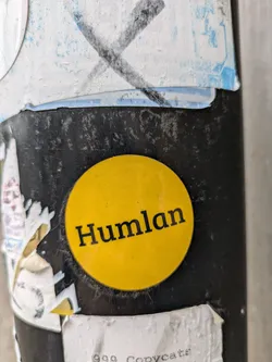 A round yellow sticker with the word "Humlan" in black text on a dark surface.  There are other stickers visible around it.