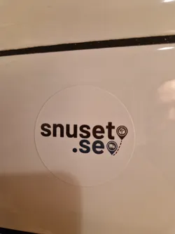 A round white sticker with the text "snuset.se" in a stylized dark font.  Small icons of a package and a delivery truck with location markers are beside the text.  The sticker appears to be affixed to a light-colored surface.