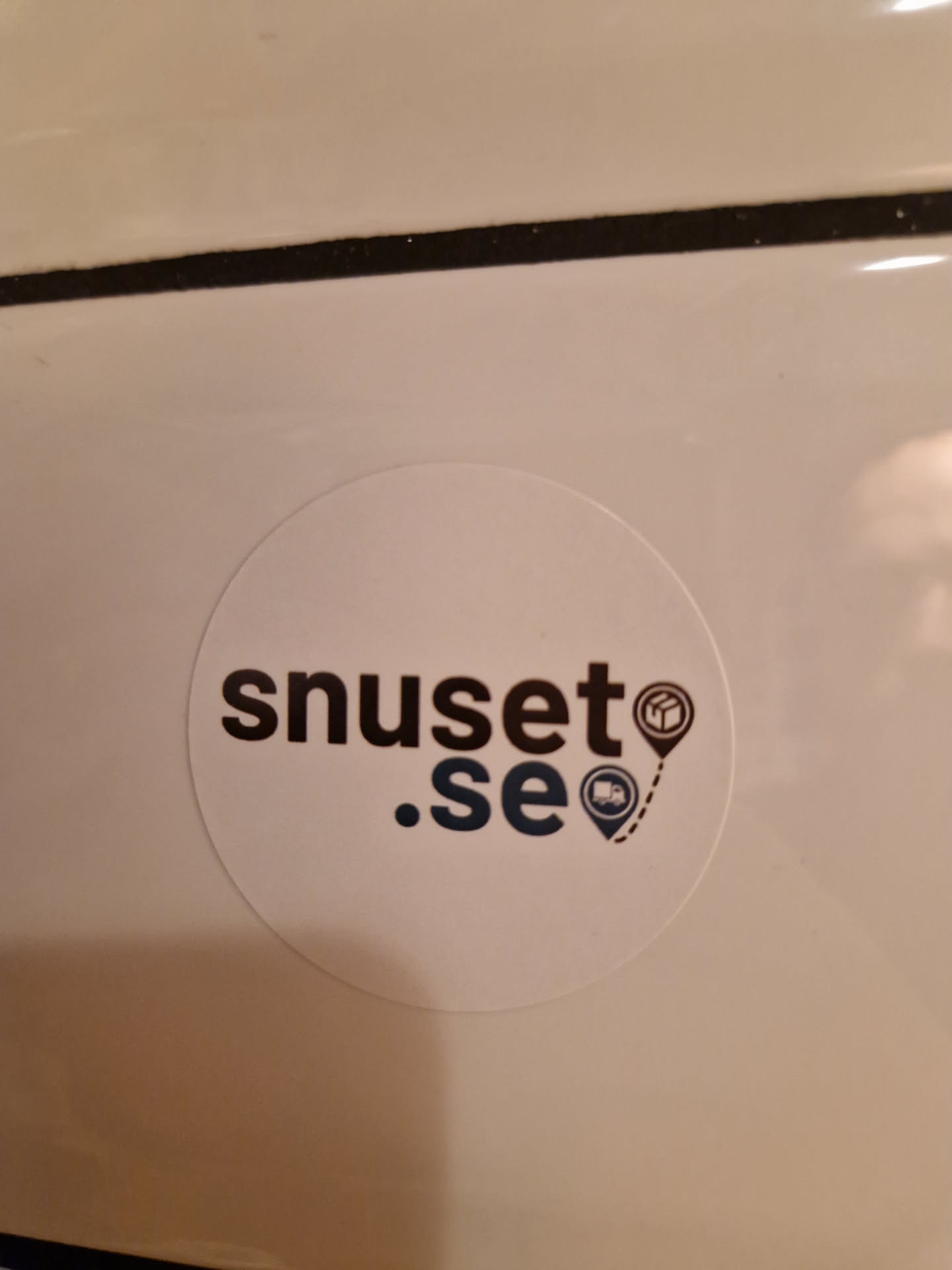 A round white sticker with the text "snuset.se" in a stylized dark font.  Small icons of a package and a delivery truck with location markers are beside the text.  The sticker appears to be affixed to a light-colored surface.