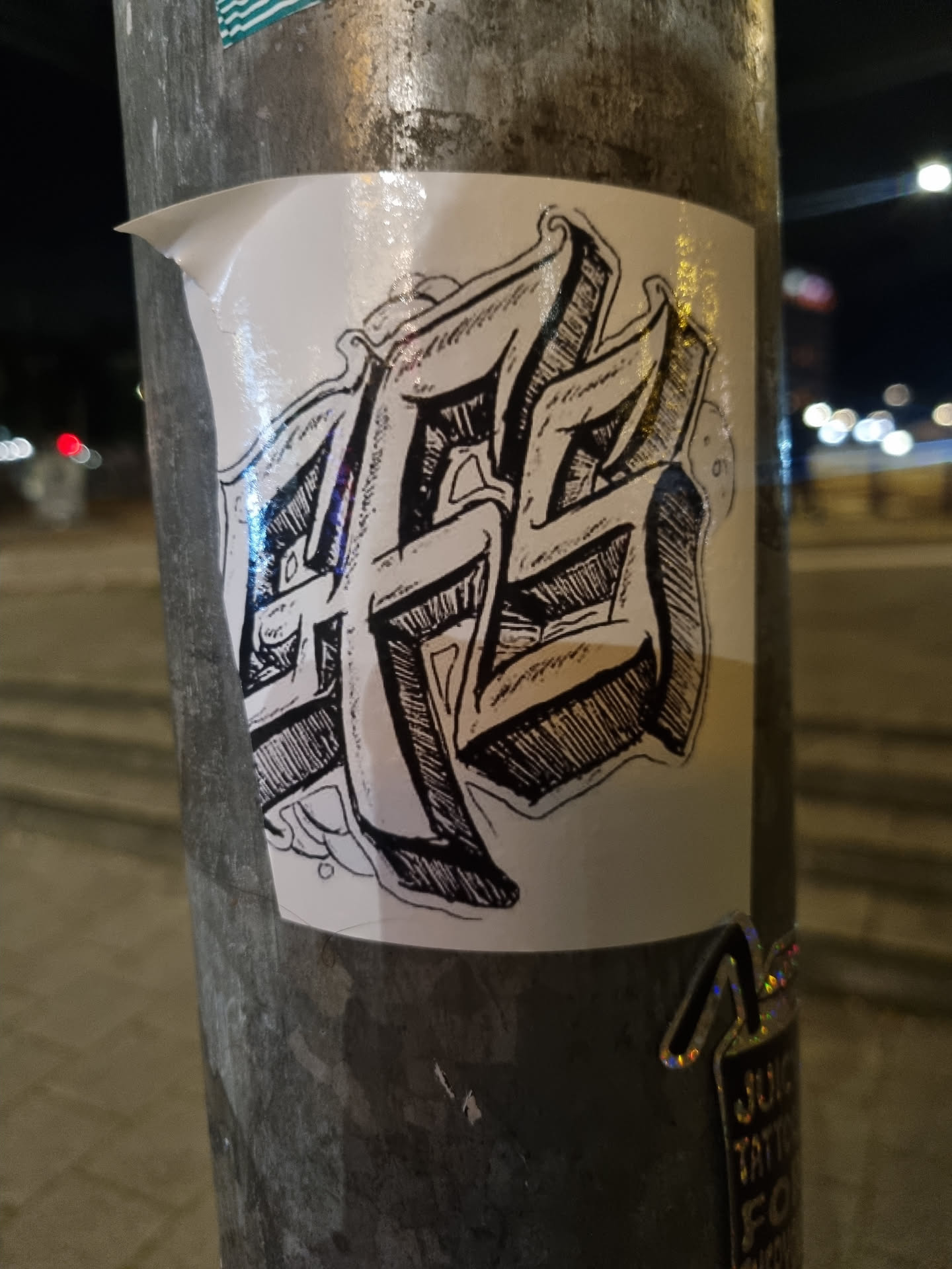 This sticker showcases a unique blend of artistic styles. The central motif is a graphic representation of stylized lettering, possibly an abbreviation or logo. The design is rendered in a bold, almost three-dimensional manner, with heavy use of shading and line work to create a sense of depth and texture. The overall aesthetic suggests a street art or graffiti influence.