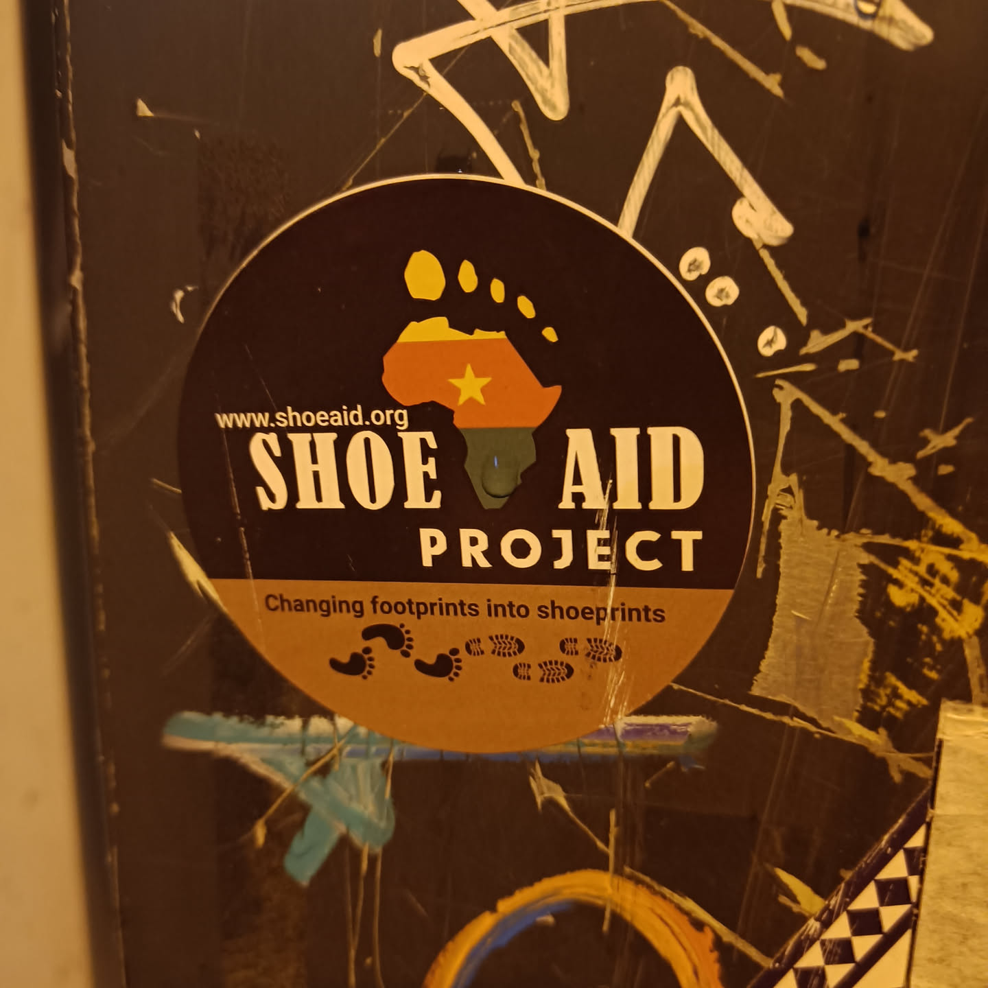 A round sticker with the Shoe Aid Project logo.  The logo features a footprint in the shape of the African continent with a yellow star, and the text 'Shoe Aid Project'.  The sticker also includes the website address www.shoeaid.org and the tagline 'Changing footprints into shoeprints'.