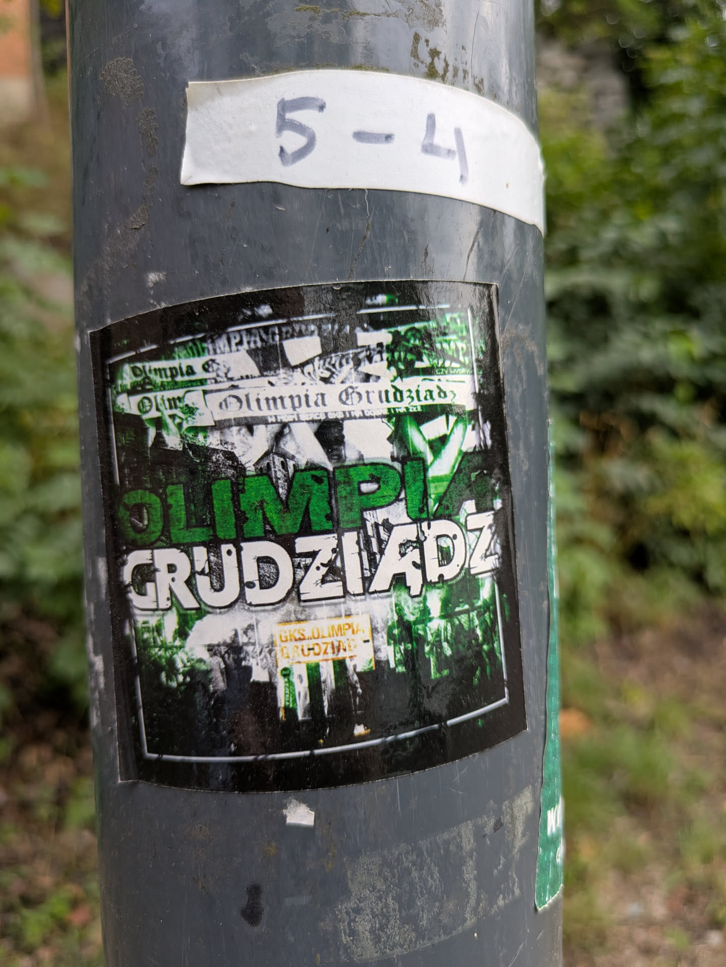 The sticker is a rectangular piece, predominantly featuring a grunge-like design in green, white, and black. The central focus is the club name "OLIMPIA" in large, vibrant green letters, stacked above "GRUDZIĄDZ" in distressed white letters with a black outline. The background is a chaotic collage of fragmented text and abstract shapes, primarily in black and white with hints of green, giving it a layered, worn appearance. In the bottom right corner, a smaller yellow-orange rectangle contains additional text. The overall aesthetic suggests a strong connection to a sports club, likely football, with a raw, fan-culture feel.