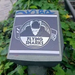 A grayscale sticker featuring a winged shark skull in a circular design with the text "FLYING SHARKS". The sticker is affixed to a gray square post surrounded by greenery.
