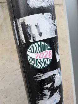 Street sticker A circular sticker slapped on a black pole, referencing Swedish politician Birgitta Ohlsson. The design features a dark green background split by a wavy white banner across the center. The top half contains the bold white text 'BIRGITTA', the central white stripe features the year '2026' printed in pink, and the bottom half contains the white text 'OHLSSON' curved along the lower edge.