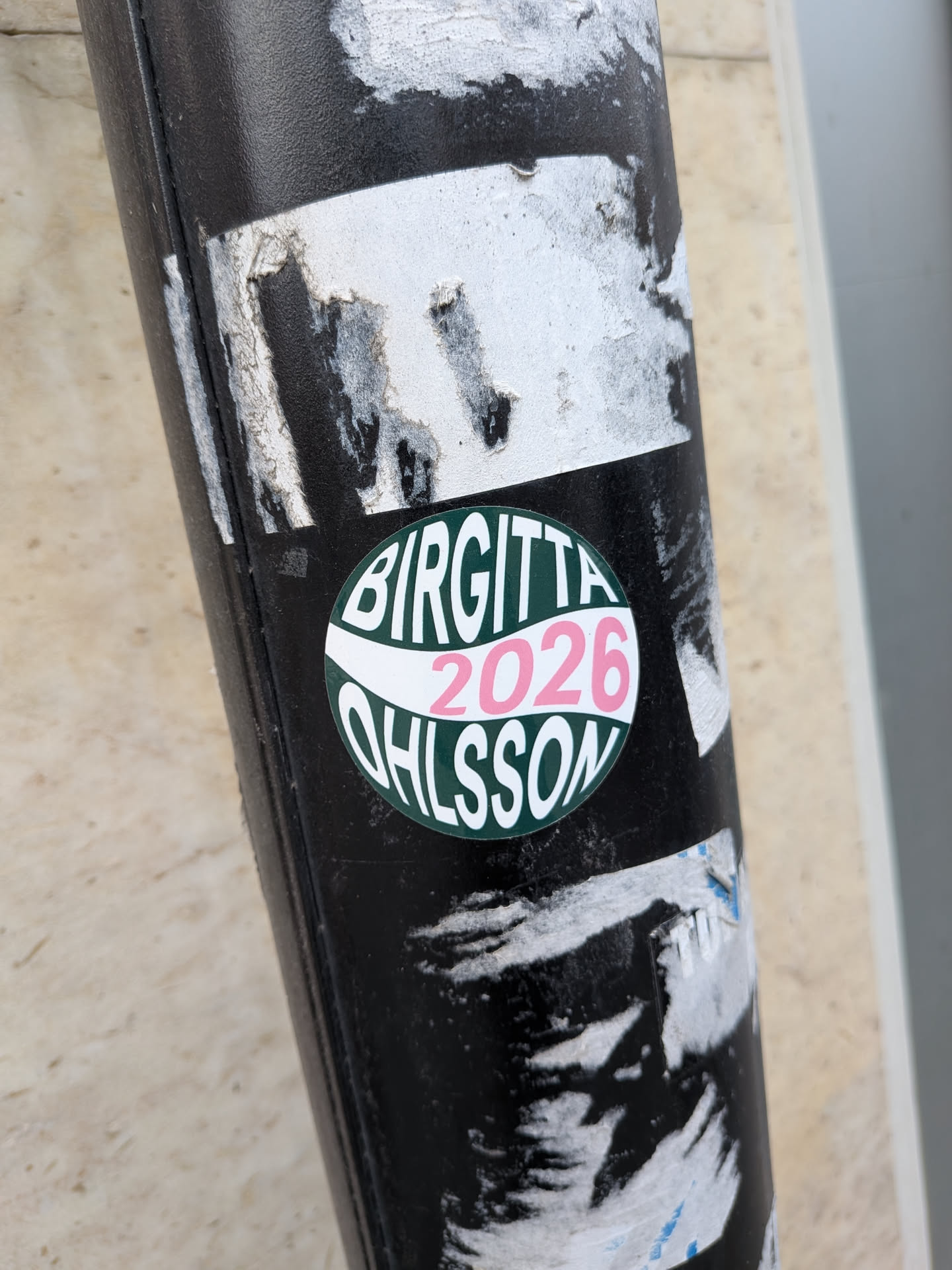 A circular sticker slapped on a black pole, referencing Swedish politician Birgitta Ohlsson. The design features a dark green background split by a wavy white banner across the center. The top half contains the bold white text 'BIRGITTA', the central white stripe features the year '2026' printed in pink, and the bottom half contains the white text 'OHLSSON' curved along the lower edge.