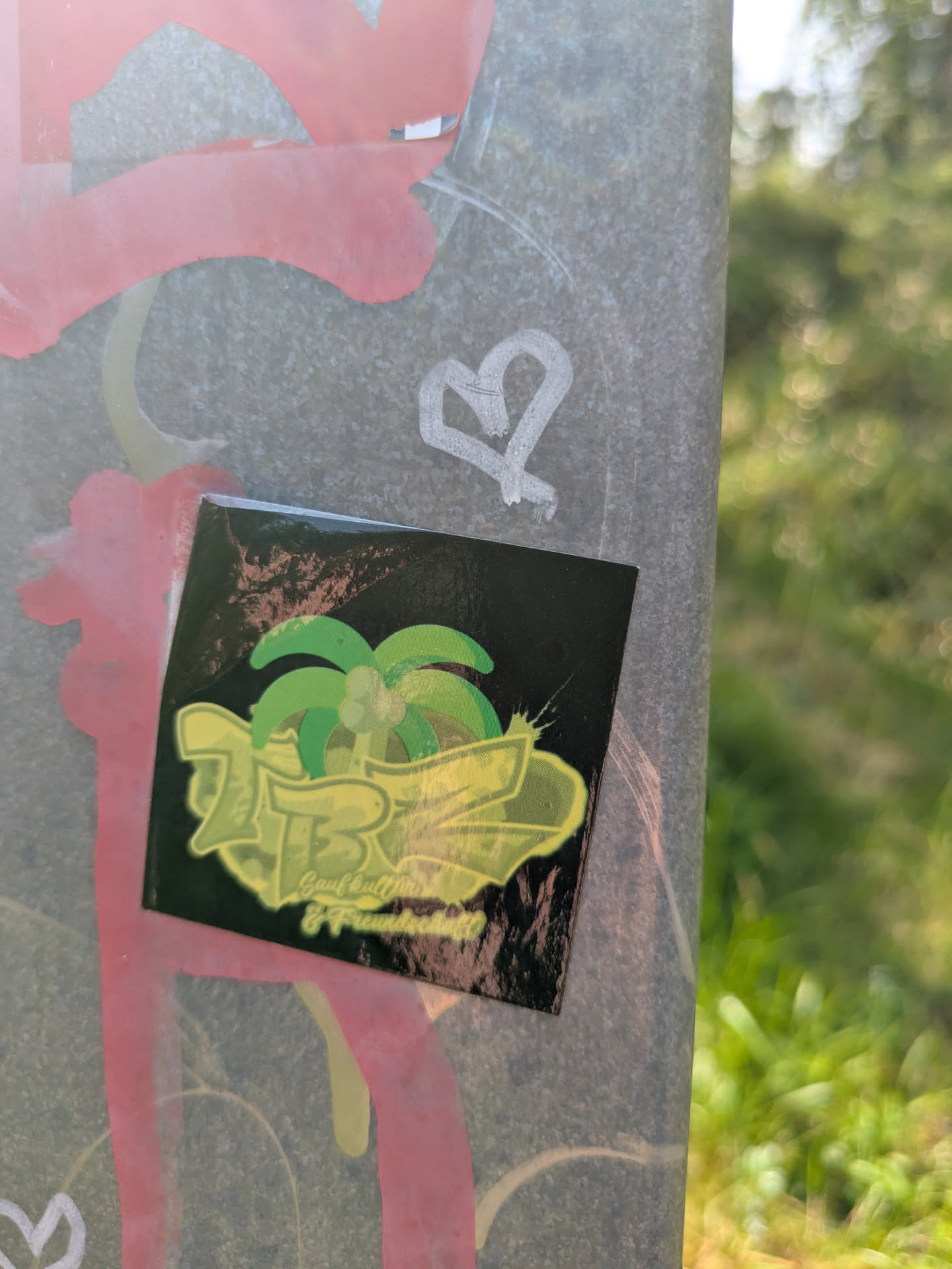 The sticker is square with a glossy finish and a black background. The central design includes a vibrant green palm tree. Overlapping and in front of the palm tree is large, stylized text that appears to read 'ZB', rendered in a bright yellow-green outline with a darker green fill, giving it a layered, almost three-dimensional effect. Beneath this large lettering, in a smaller, lighter font, the German words 'Gaufkultur & Freundschaft' are visible. The sticker is placed on a textured grey pole or wall, which also bears pink and white graffiti marks, including a heart symbol.