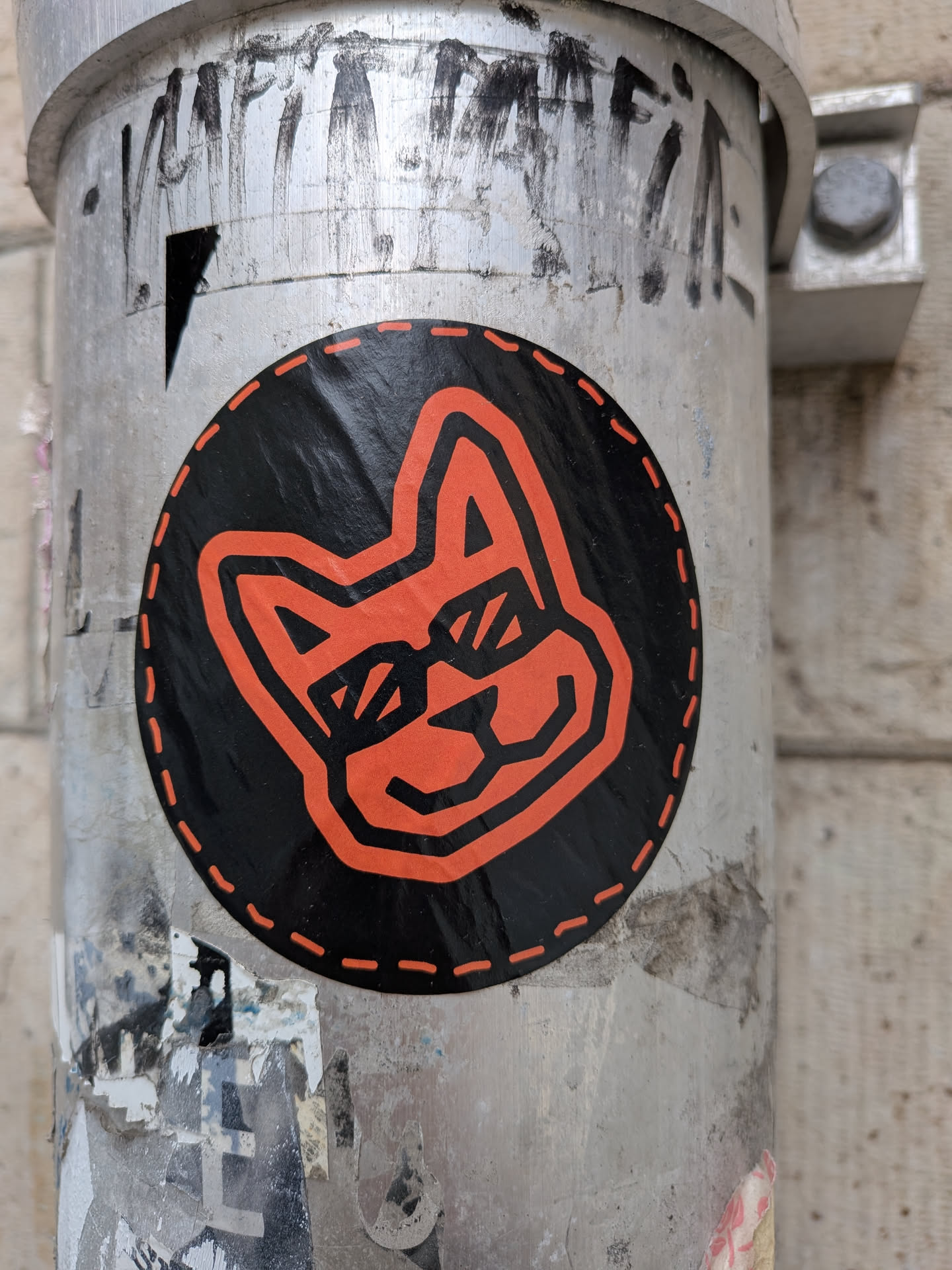 A circular sticker featuring a stylized, minimalist head of an animal, likely a fox or dog, rendered in bright orange against a solid black background. The animal has pointed ears, a snout, and its eyes are depicted as rectangular sunglasses. The entire design is outlined with bold, geometric lines. A distinctive orange dashed line, resembling stitching, encircles the perimeter of the black circle. The sticker has a slightly wrinkled texture, indicating it's adhered to a surface.