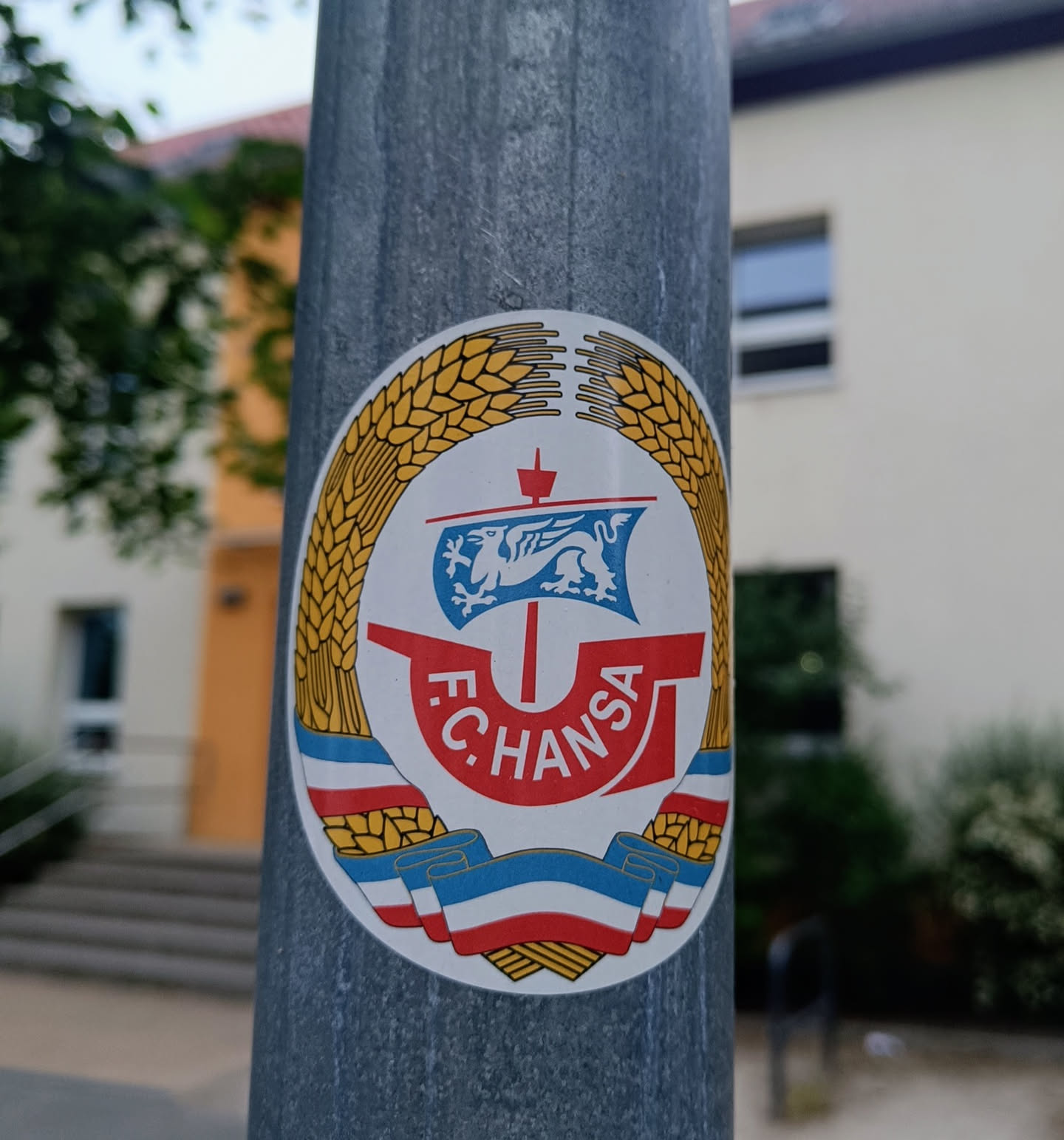 A round sticker featuring the logo of FC Hansa Rostock, a German football club. The logo includes a ship with a griffin, surrounded by wheat and ribbons in the colors of the club.  The sticker is affixed to a gray metal lamppost.