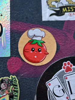 A round sticker featuring a cartoon tomato wearing a chef's hat. The tomato has a happy expression and a small plus sign on its body. The background is a light beige color.