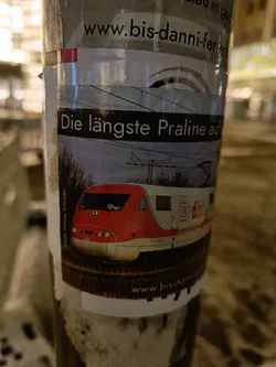 Sticker on a pole advertising www.bis-danni-ferrero.de, featuring an image of a high-speed train.  The text on the sticker says "Die längste Praline auf" which translates to "The longest chocolate bar on".