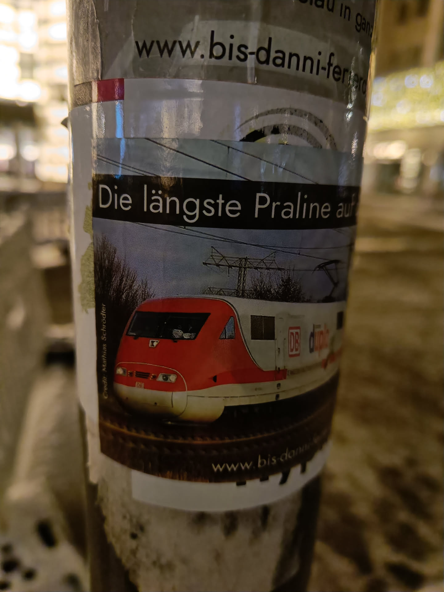 Sticker on a pole advertising www.bis-danni-ferrero.de, featuring an image of a high-speed train.  The text on the sticker says "Die längste Praline auf" which translates to "The longest chocolate bar on".