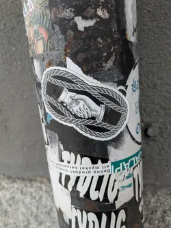 Close-up of a sticker on a pole. The sticker shows two hands shaking, encircled by a thick rope. The style is simple line art with black ink on a white background. The sticker shows some minor wear.