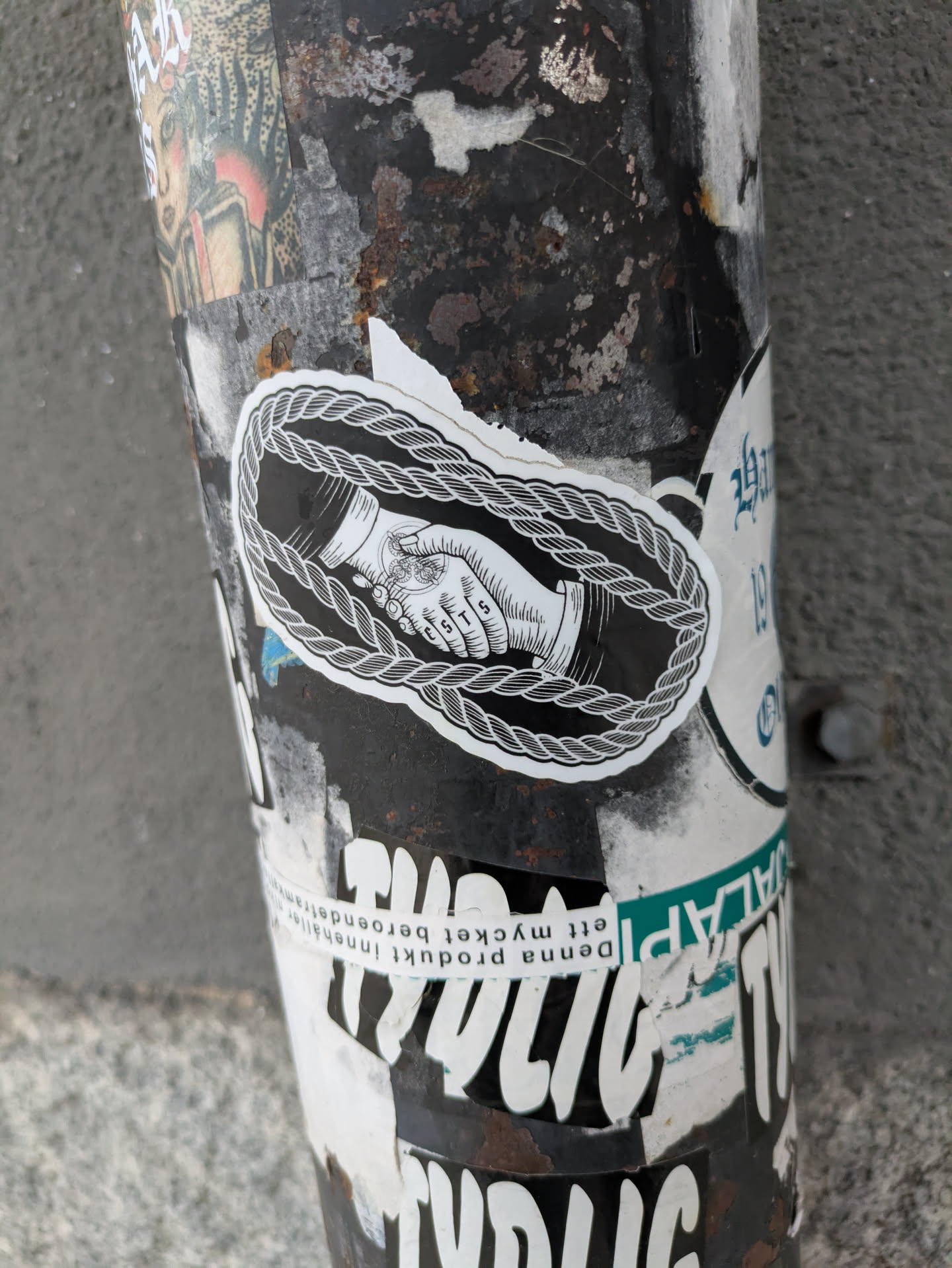 Close-up of a sticker on a pole. The sticker shows two hands shaking, encircled by a thick rope. The style is simple line art with black ink on a white background. The sticker shows some minor wear.