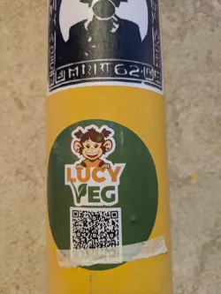 This sticker features a smiling cartoon monkey with pigtails, wearing a shirt, above the words "LUCY VEG" in orange and green. The "E" in VEG is stylized with a leaf. Beneath the text is a QR code. The sticker is on a yellow background.