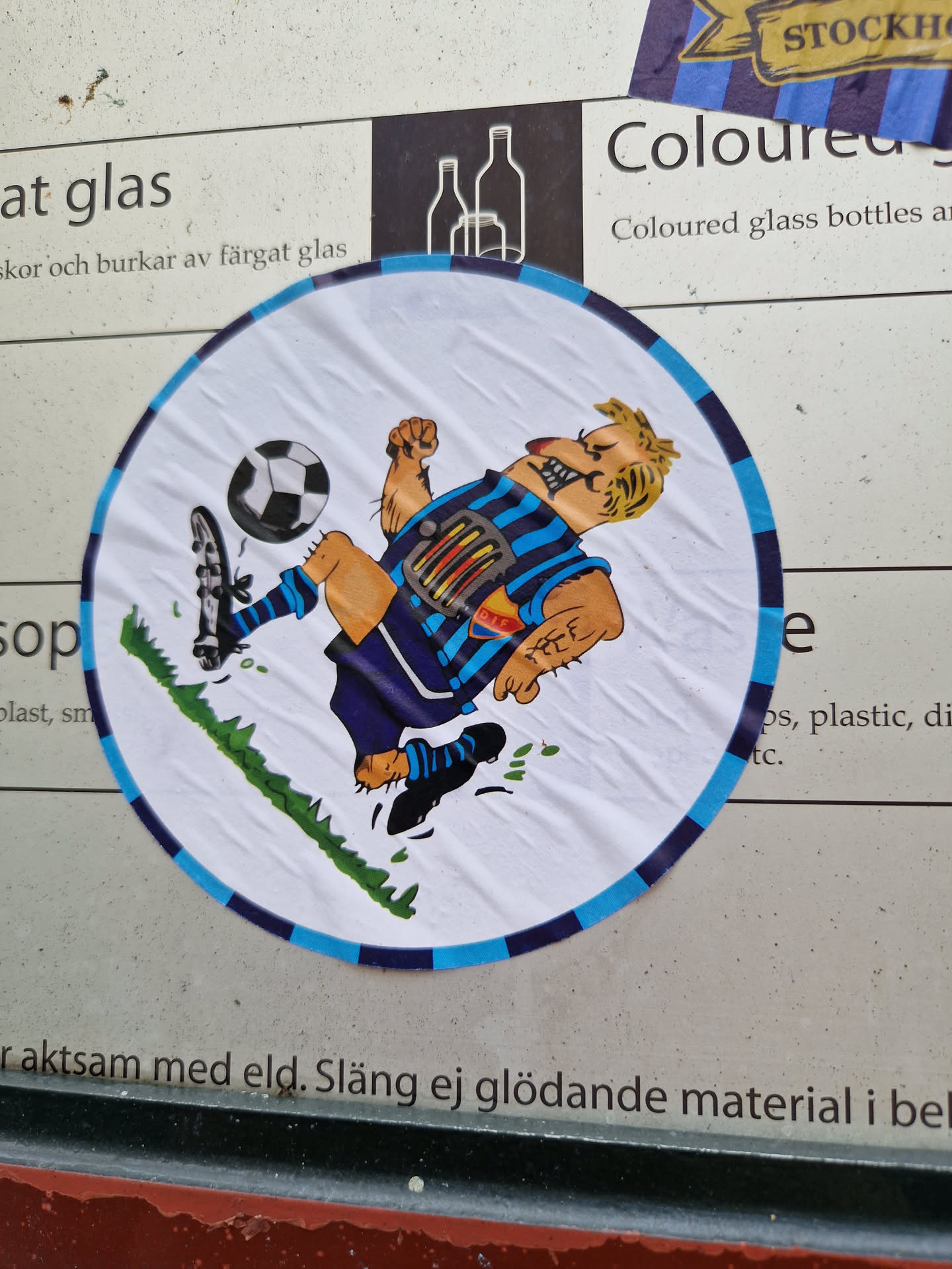 A round sticker featuring a cartoon illustration of a man playing soccer. He has light skin, blond hair, and is wearing a blue and white soccer uniform. He is kicking a soccer ball with a determined expression on his face. The background of the sticker is white with a touch of green grass.  The sticker has a dark blue border.