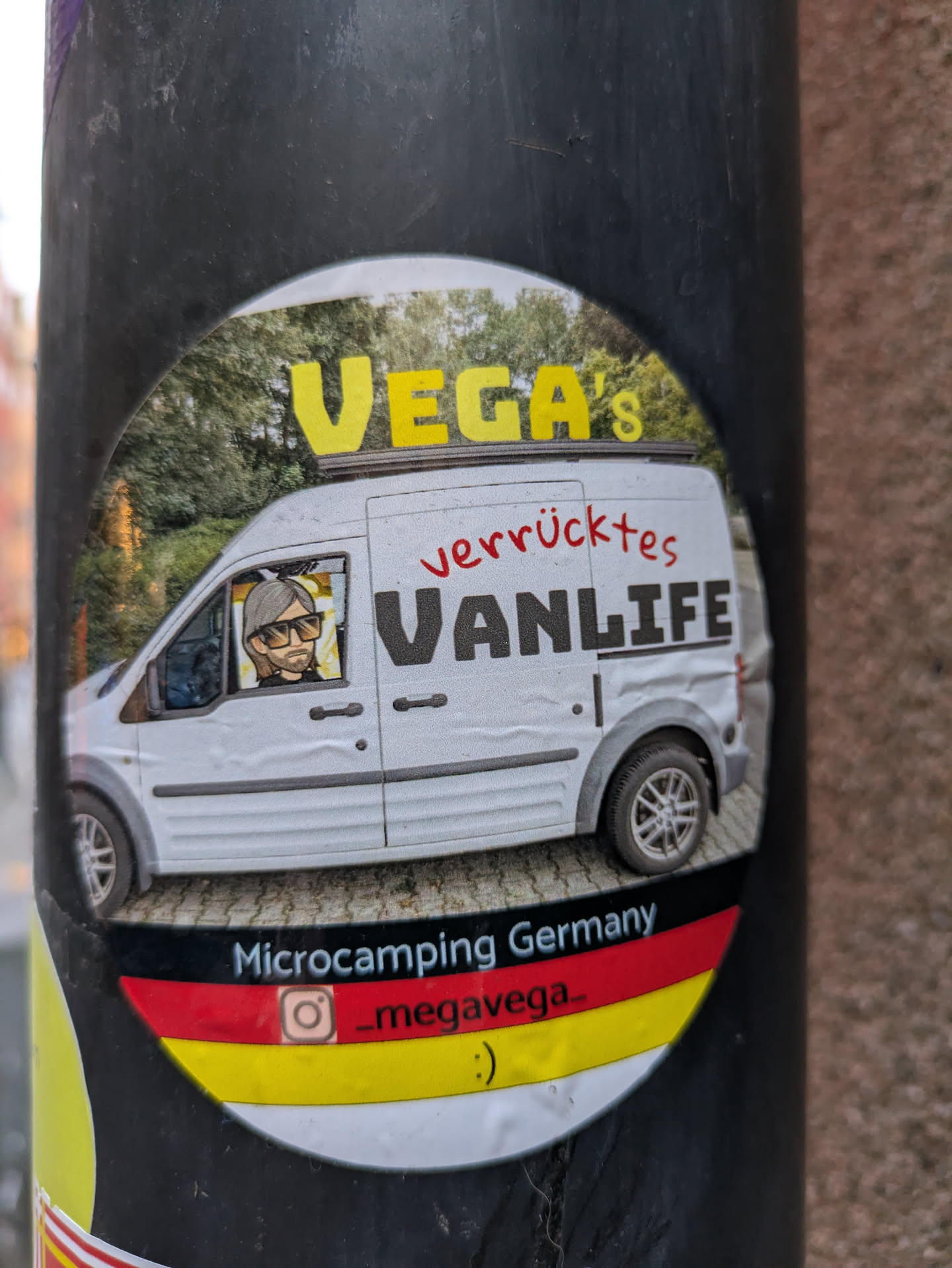 A round sticker featuring an illustration of a white van with a person inside. The text on the sticker says "VEGA's verrücktes VANLIFE" and "Microcamping Germany". There's an Instagram handle and a smiley face at the bottom. The sticker uses the colors of the German flag.