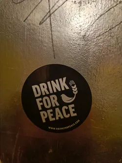 A round black sticker with the text "Drink for Peace" in white, along with an image of a bird and wheat.  A website address is included at the bottom. The sticker is affixed to a textured, gold-colored surface.