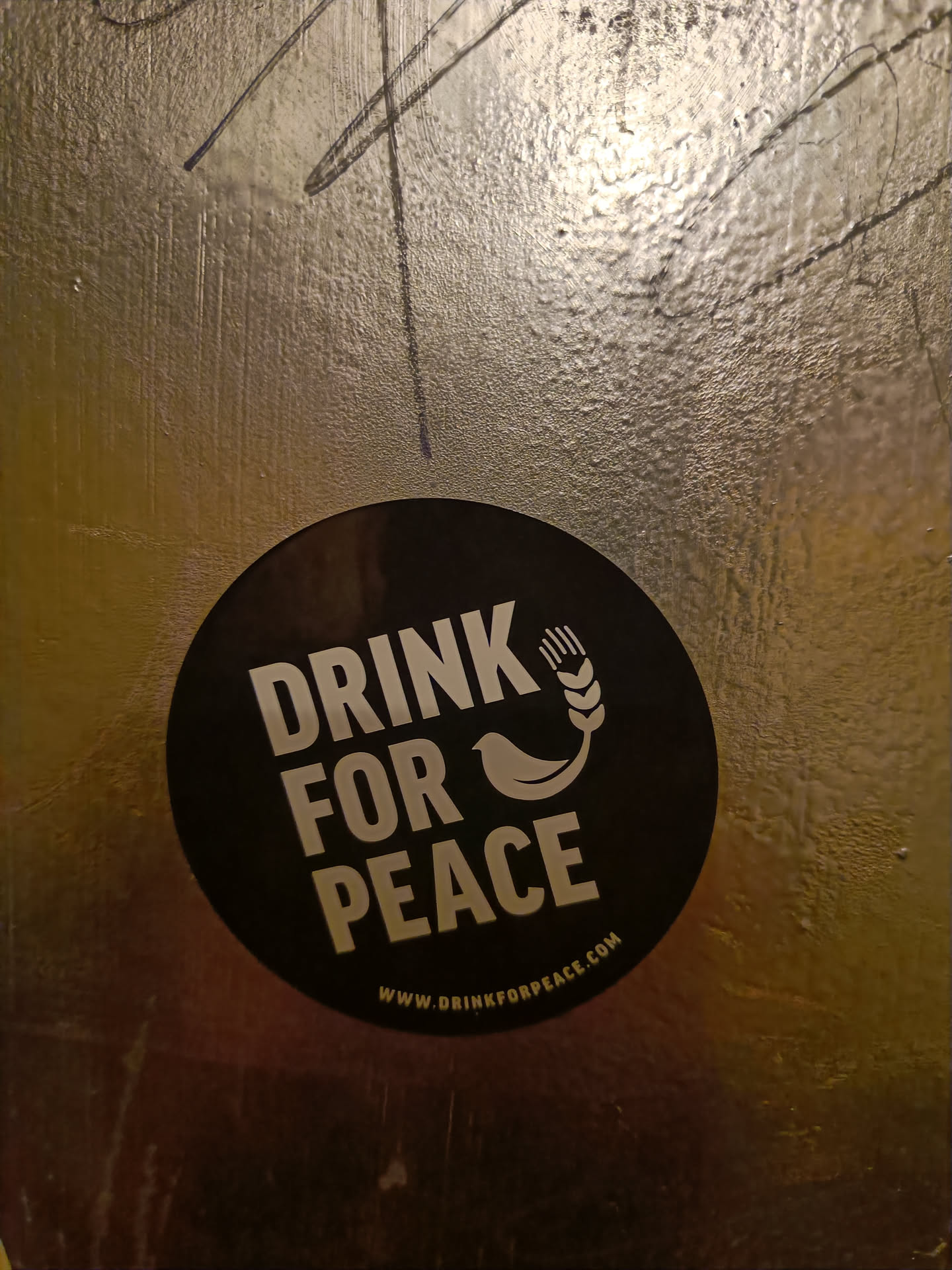 A round black sticker with the text "Drink for Peace" in white, along with an image of a bird and wheat.  A website address is included at the bottom. The sticker is affixed to a textured, gold-colored surface.