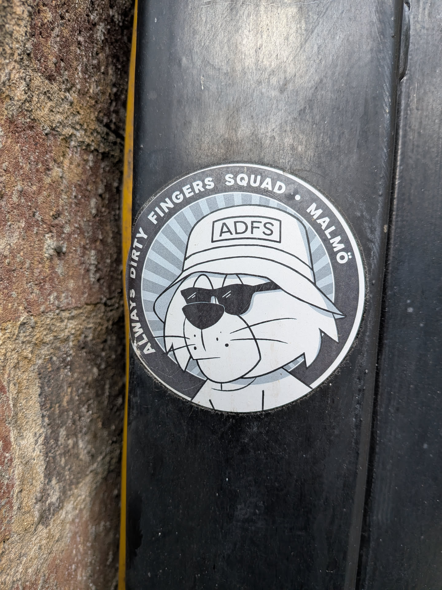 A circular sticker featuring a cartoon cat's head and upper torso. The cat is depicted in a grayscale palette with white fur and black outlines. It wears dark, rectangular sunglasses and a white bucket hat with the letters 'ADFS' prominently displayed on the front. The background behind the cat consists of radiating light grey lines, creating a sunburst effect. A dark grey outer ring encircles the central design, containing white text. The sticker has a slightly worn appearance, indicating it's a physical object adhered to a surface.