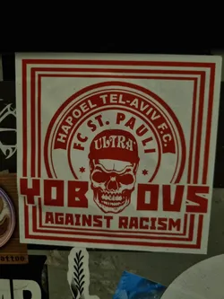 Red and white sticker with a skull graphic and text. It features the names of two football clubs, Hapoel Tel Aviv and FC St. Pauli, along with the word "Ultras" and the phrase "Against Racism". The skull is wearing a cap, and stars are placed on either side.