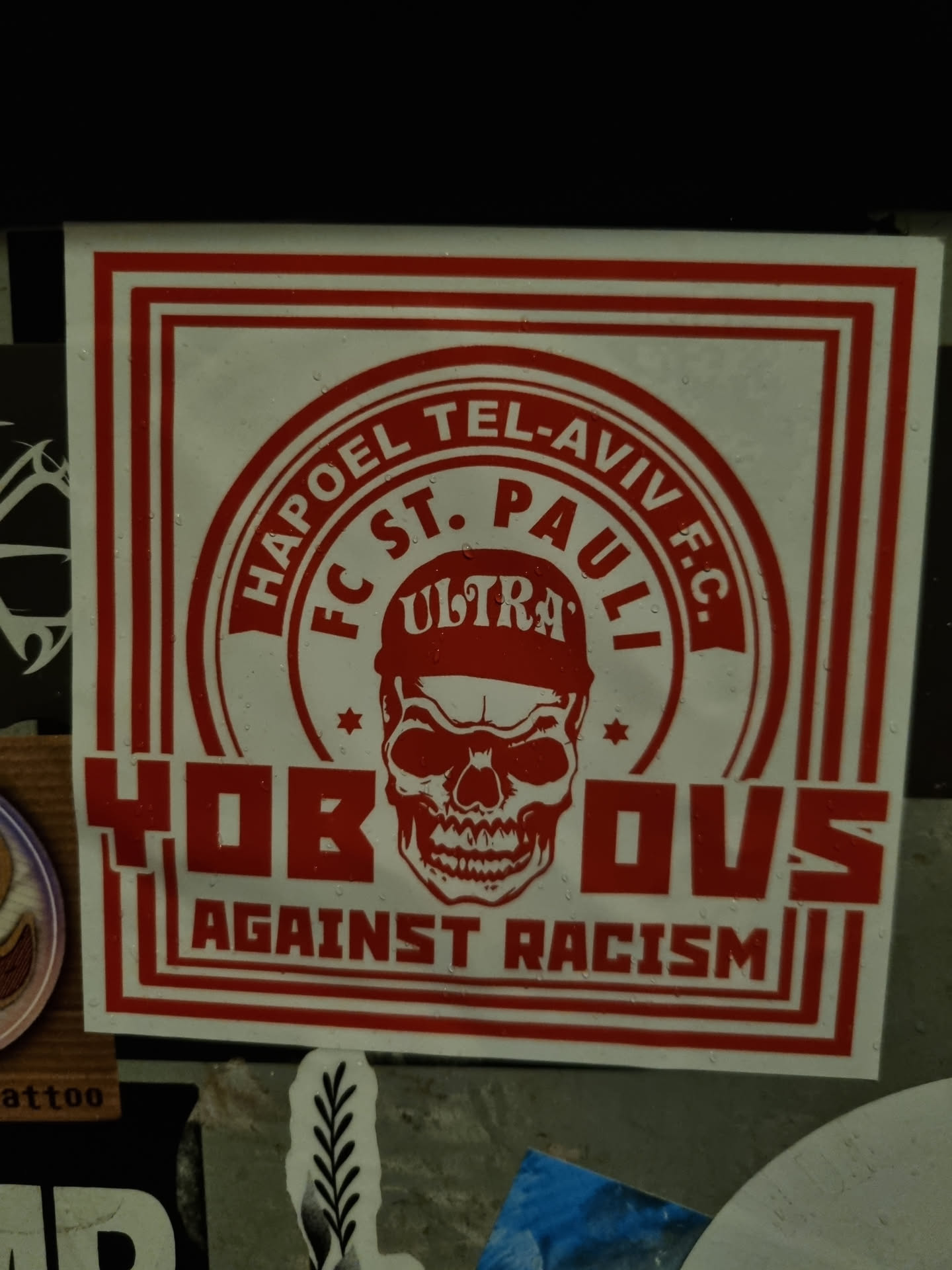 Red and white sticker with a skull graphic and text. It features the names of two football clubs, Hapoel Tel Aviv and FC St. Pauli, along with the word "Ultras" and the phrase "Against Racism". The skull is wearing a cap, and stars are placed on either side.