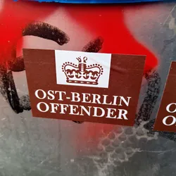 A brown sticker with a crown and the text "OST-BERLIN OFFENDER" in white. The sticker is affixed to a surface with red graffiti.