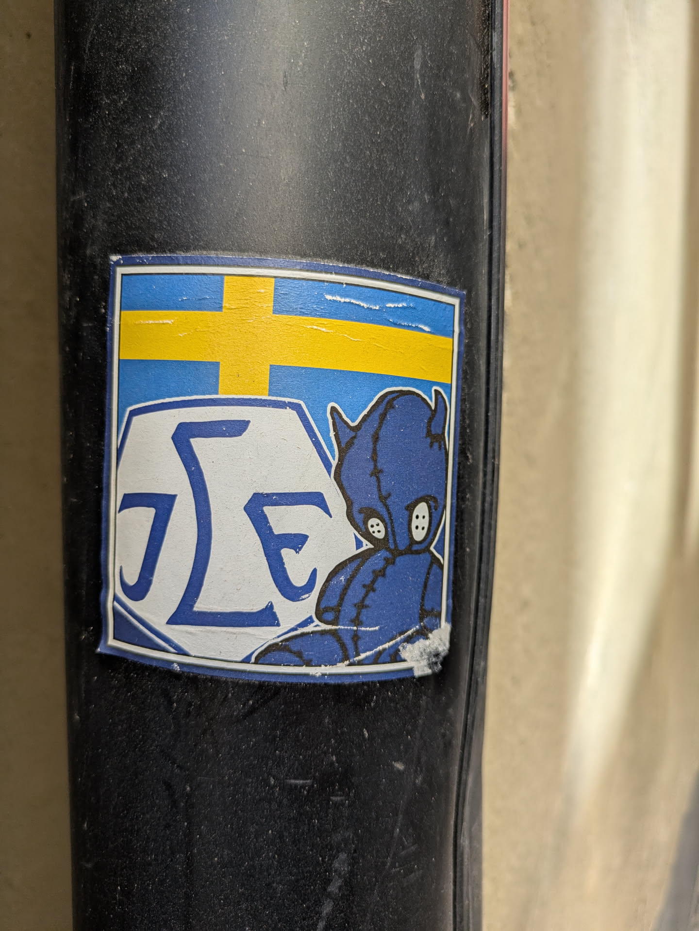 The sticker has a square shape with a blue and yellow Swedish flag design at the top. Below the flag, there is a white area with the letters 'JLE' stacked on top of each other, designed in blue. To the right of the letters, there is a blue teddy bear character. The sticker is affixed to a dark, possibly rubber surface.