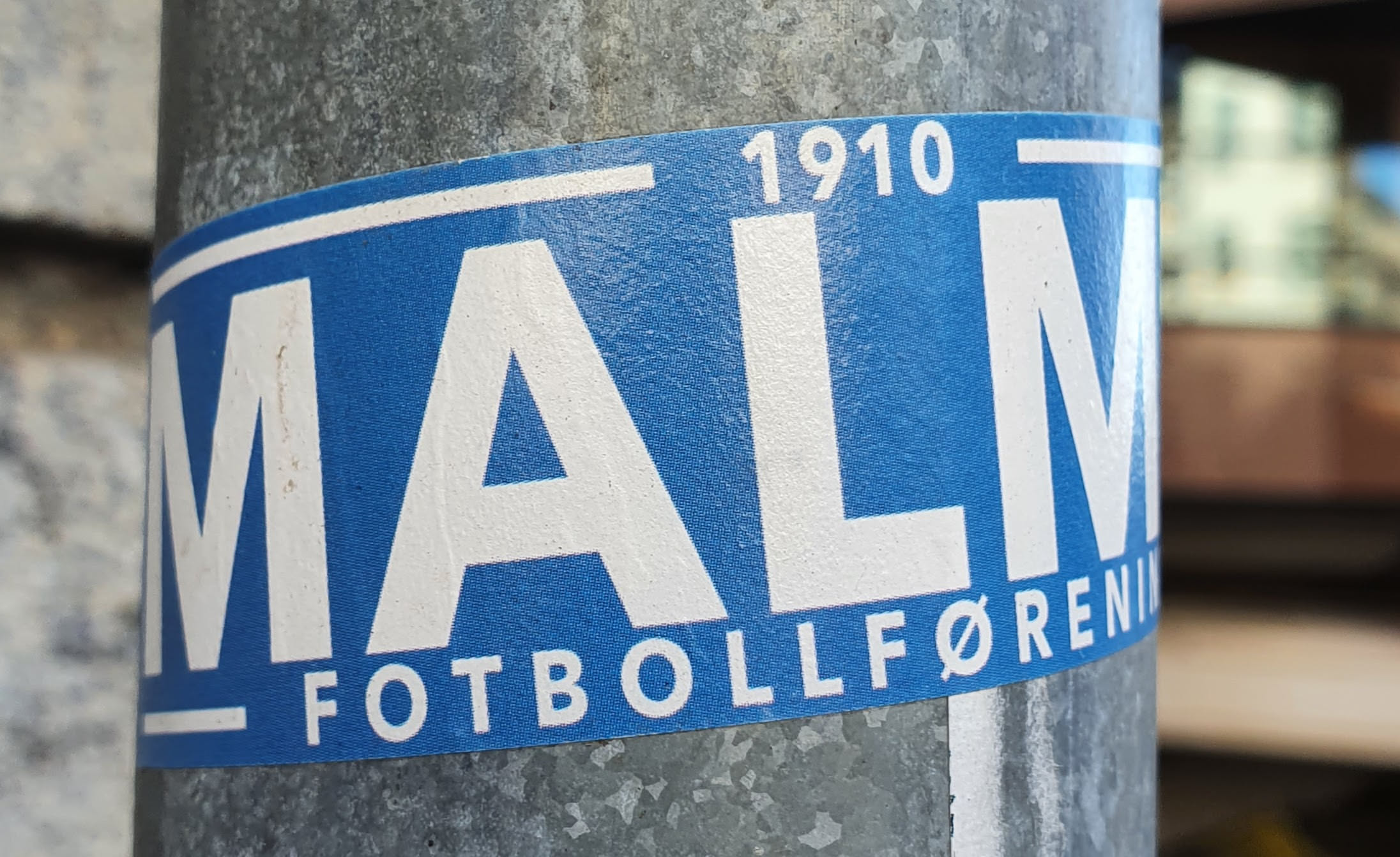 A blue and white sticker on a metal pole.  The sticker features the text MALMÖ in large white lettering on a blue background, with the smaller text "1910" and "FOTBOLLFÖRENING" visible.