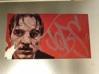 This rectangular sticker showcases a close-up portrait of a man with intense eyes and a mustache. The background is a vibrant red, with a stylized grey signature or tag prominently placed over it.  The sticker shows some minor wear and tear at the edges.