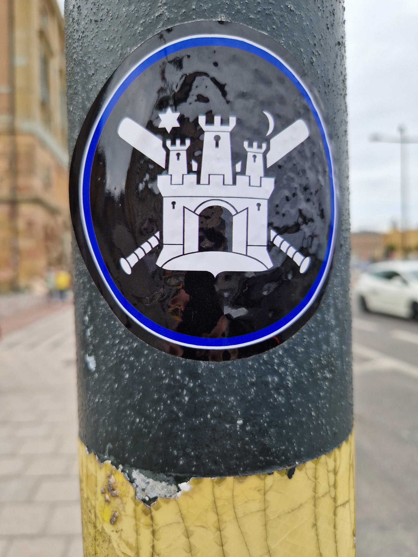 This round sticker displays a black and white illustration of a castle with crenelations, a gate, and two towers.  The castle is centrally positioned, with a baseball bat crossing over another on each side.  Above the castle, there's a six-pointed star to the left and a crescent moon to the right. The sticker has a black background with a blue outer ring.