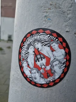 A round sticker featuring a black and white illustration of several expressive faces and figures, arranged in a circular composition.  The style is reminiscent of vintage printmaking. The background is a red and black checkered pattern.  The text at the top of the circular illustration appears to read "hast du in mein Glas getan."  There is a word at the bottom that may be a signature or artist name, partially obscured but appears to read "SALTIAR".