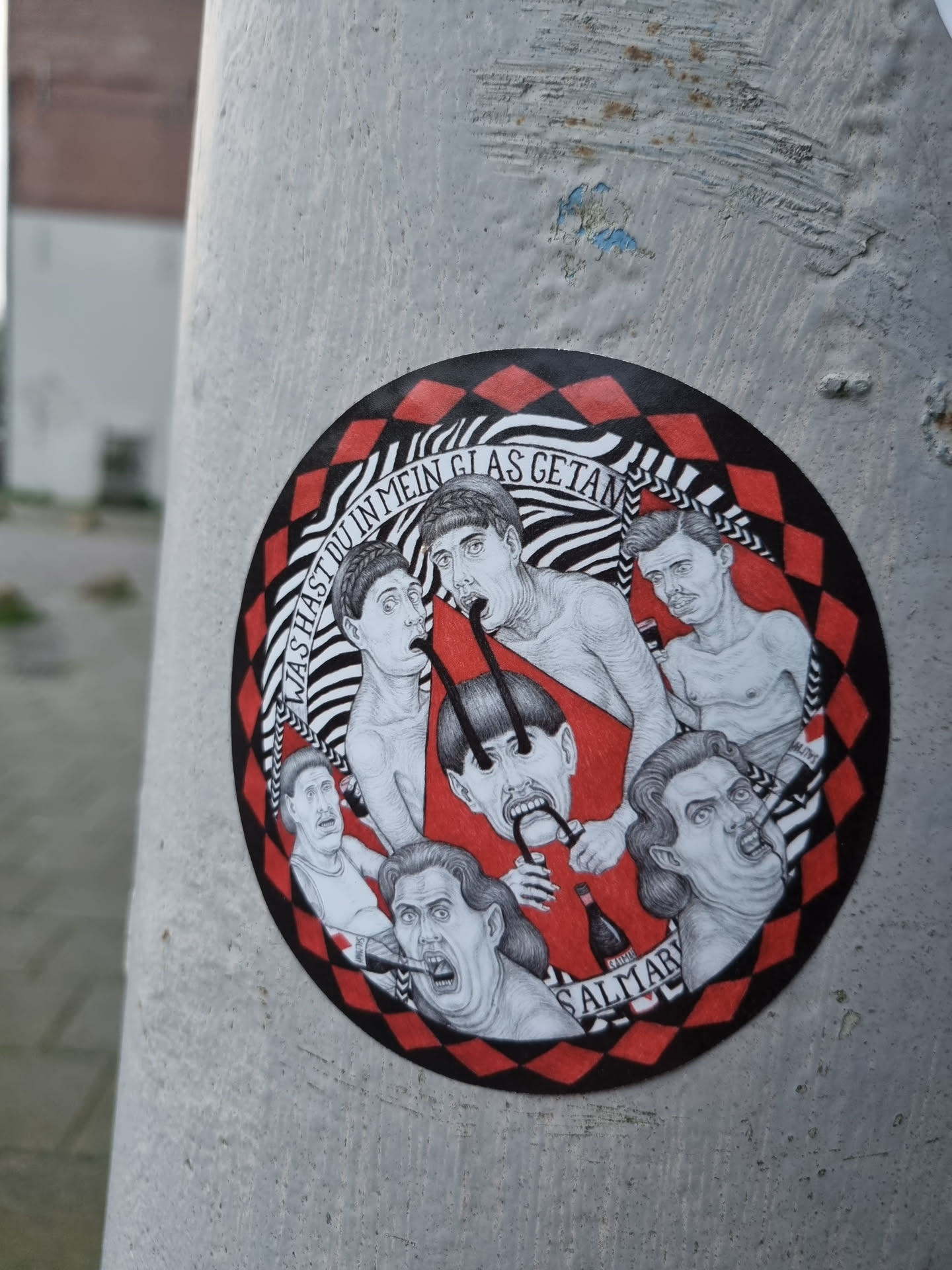 A round sticker featuring a black and white illustration of several expressive faces and figures, arranged in a circular composition.  The style is reminiscent of vintage printmaking. The background is a red and black checkered pattern.  The text at the top of the circular illustration appears to read "hast du in mein Glas getan."  There is a word at the bottom that may be a signature or artist name, partially obscured but appears to read "SALTIAR".