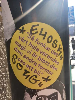 A yellow circular sticker with text in Swedish. The text discusses the interconnectedness of individuals and how they shape each other's existence.  There are additional markings around the main text.