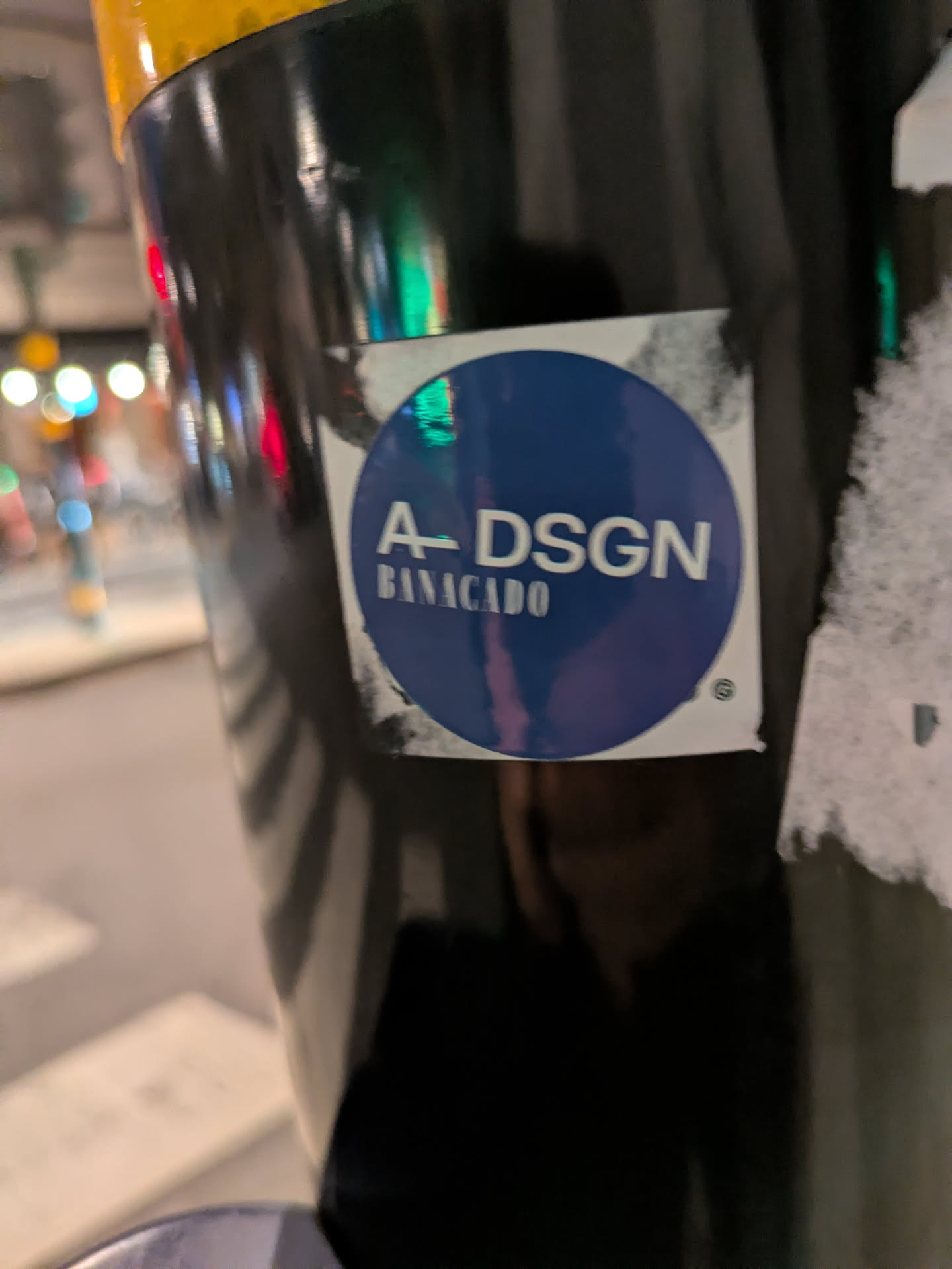 The sticker features a prominent dark blue circle centered on a white square background. The white edges of the square show signs of wear, dirt, and slight peeling. Inside the blue circle, the main text "A-DSGN" is displayed in white, sans-serif capital letters, with a horizontal line connecting the 'A' and 'D'. Below this, in smaller white capital letters, is the word "BANACADO". A small registered trademark symbol (®) is visible near the bottom right edge of the blue circle. The sticker is affixed to a reflective, dark cylindrical object, likely a pole, which shows blurred reflections of city lights in the background.