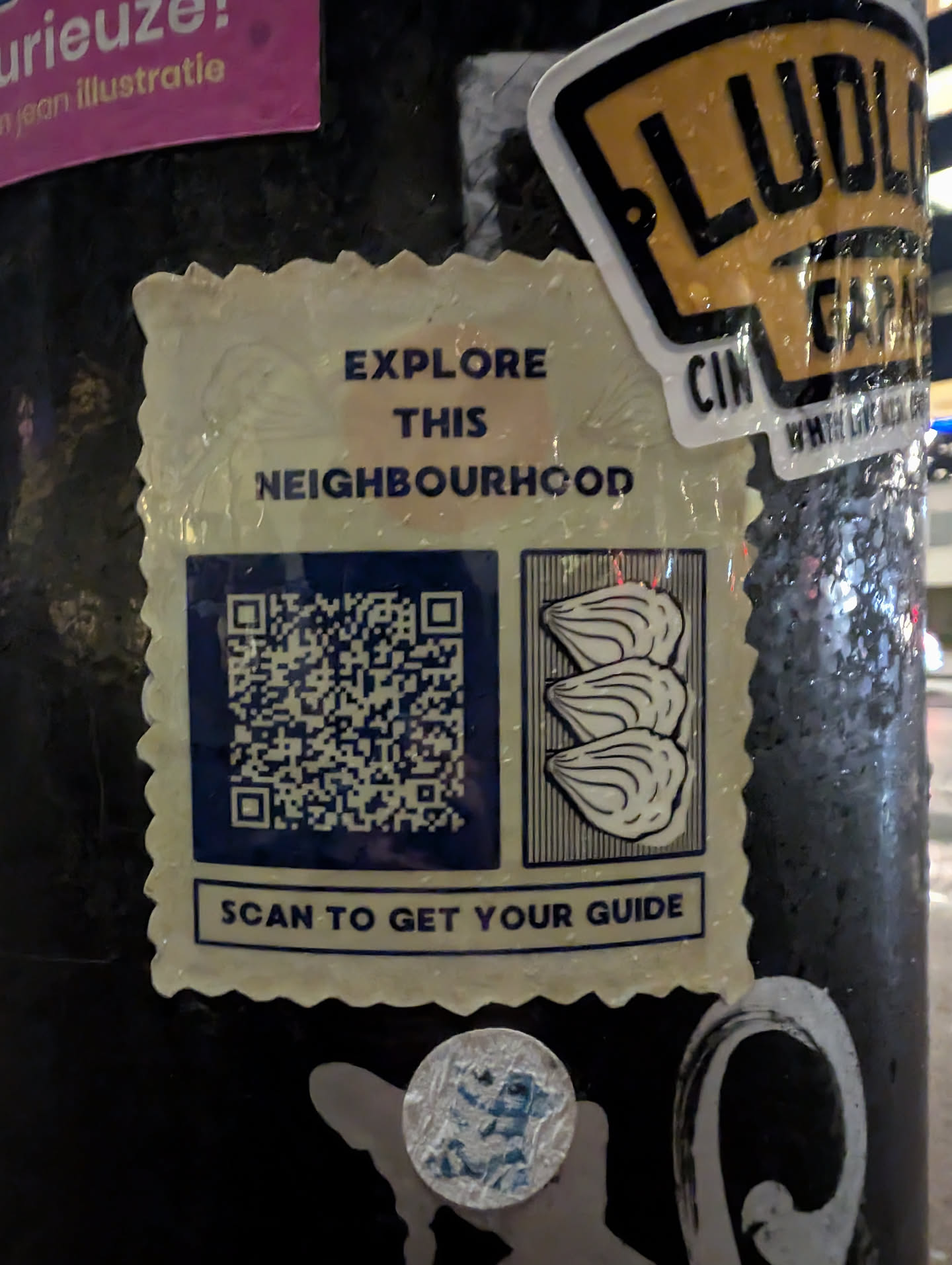 A sticker with a QR code inviting people to scan it to get a neighbourhood guide. The sticker also features an illustration of three scallops or similar shellfish.