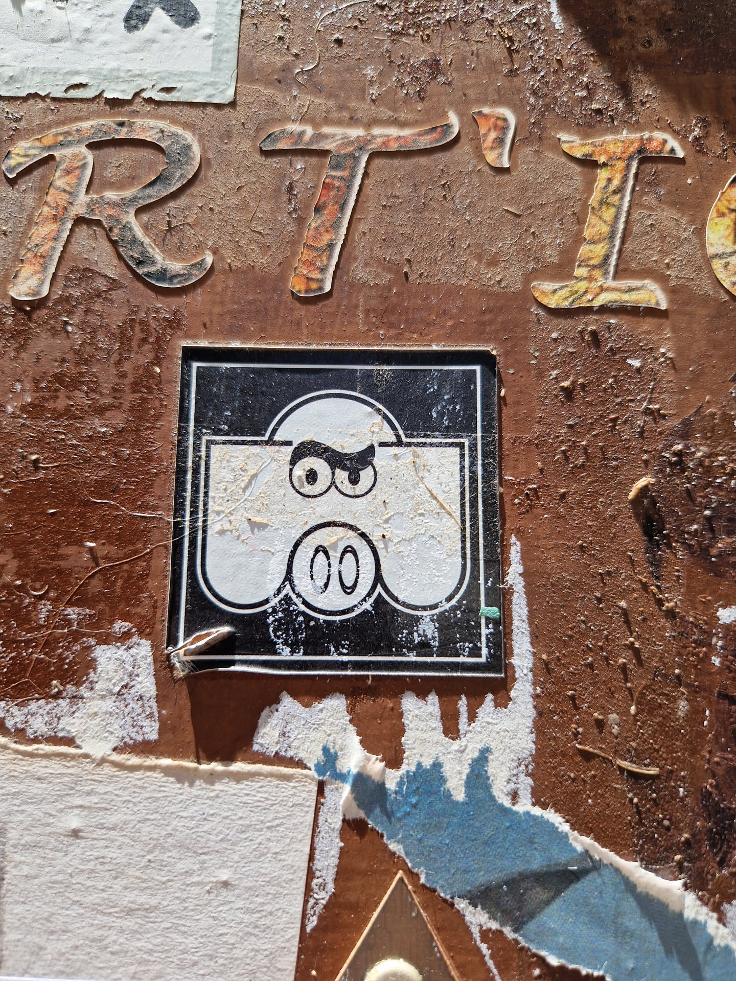 A square sticker featuring a simple cartoon illustration of a character with angry eyebrows and a small mouth. The sticker has a black background with a white character in the center and a number 00 at the bottom. The sticker is pasted on a rusty brown wall covered in other stickers and graffiti.