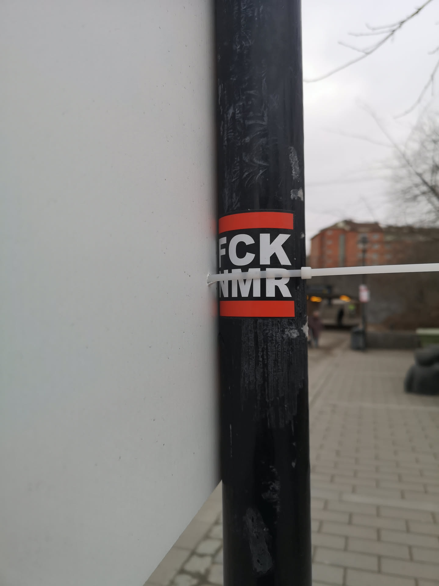 The sticker is rectangular, featuring a black background with thick red borders at the top and bottom. White, sans-serif capital letters spell out 'FCK' on the upper black section and 'NMR' on the lower black section. The sticker is affixed to a textured black pole, with a white zip tie passing over its center, securing a white board or sign to the pole. The sticker itself appears slightly worn or creased in places.
