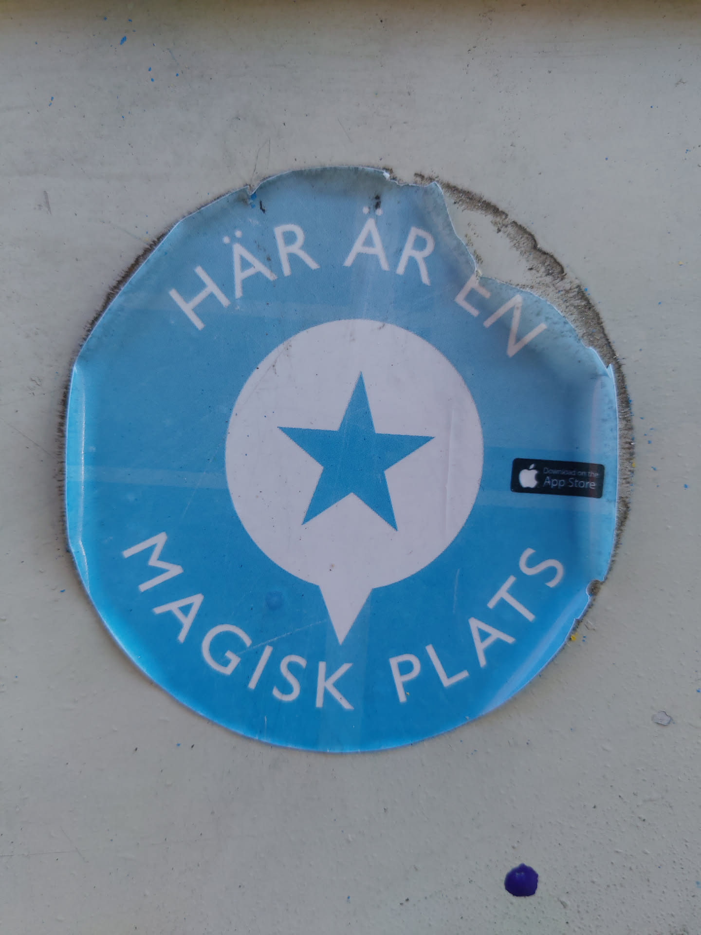 A round blue sticker with a white speech bubble containing a blue star. The text around the circle says "HÄR ÄREN MAGISK PLATS" which translates to "Here is a magical place" in Swedish. There is also an App Store logo on the sticker. The sticker is slightly damaged around the edges.