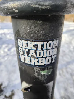 The image shows a sticker affixed to a black metal pole. The sticker reads "SEKTION STADION VERBOT" in bold white letters with a blue outline. The pole is weathered with some chipped paint and surface wear. The background is blurred and shows a snowy or icy landscape.