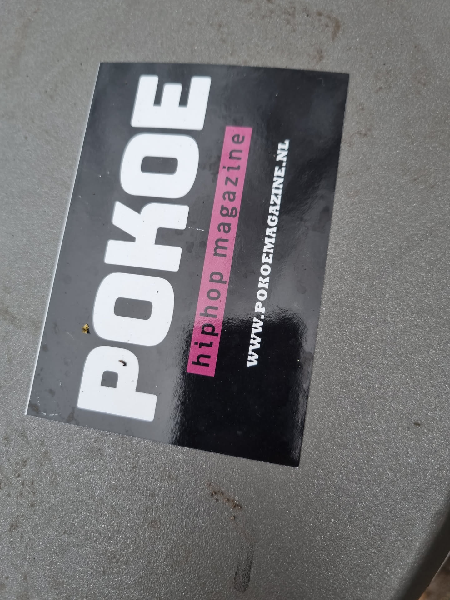 A black and white sticker with the text POKOE in large white letters,  the words "hiphop magazine" written in smaller pink letters and website address www.pokoemagazine.nl at the bottom. 